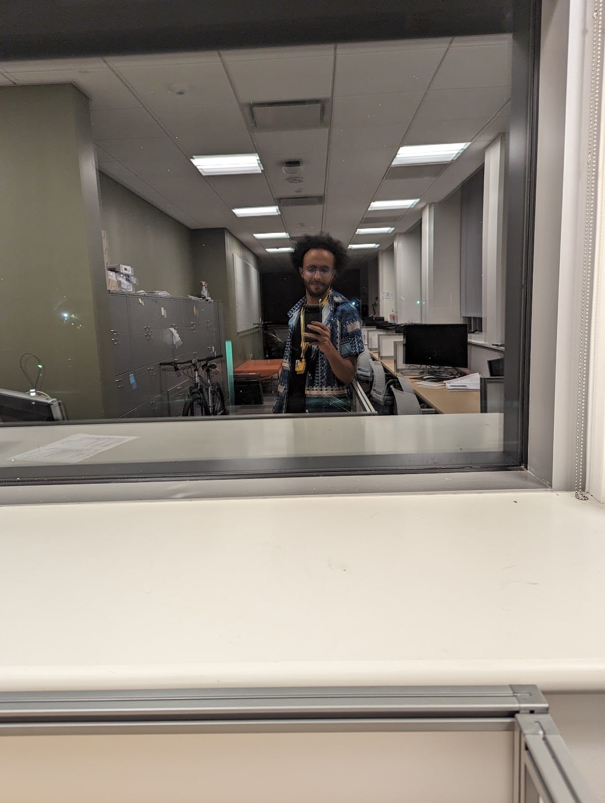 A reflection in the glass wall of an empty office at night. The author stands alone among rows of dark monitors and vacant desks, phone raised, the corridor lights running away behind him.