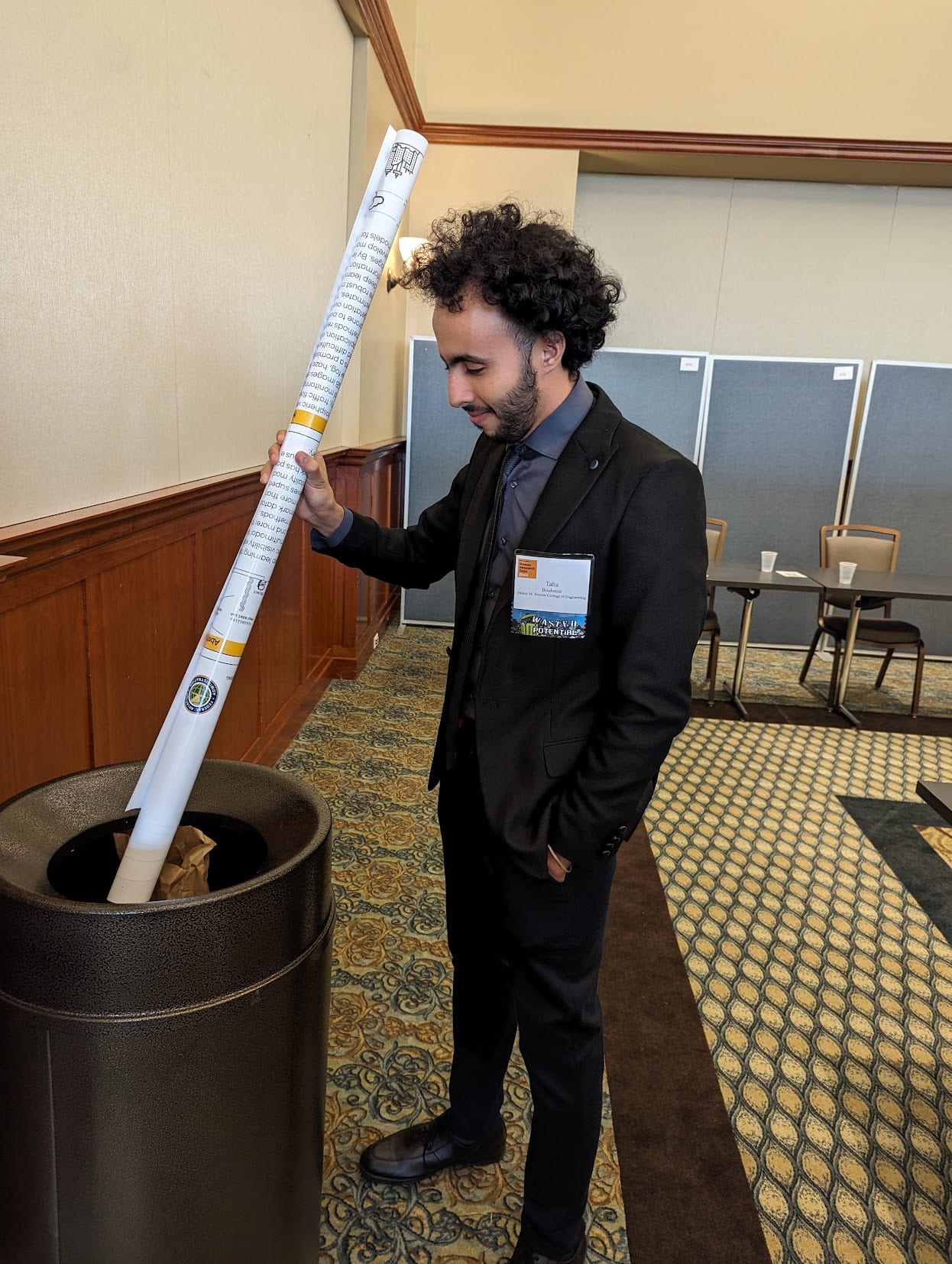 A man in a suit at a research symposium dropping a rolled-up research poster into a trash can. His conference badge reads WASTED POTENTIAL.